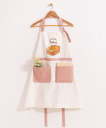 White and dusty pink apron hangs on a clothing hanger. A bundle of flowers and a notebook are placed in the oversized pink pockets on the front of the apron.
