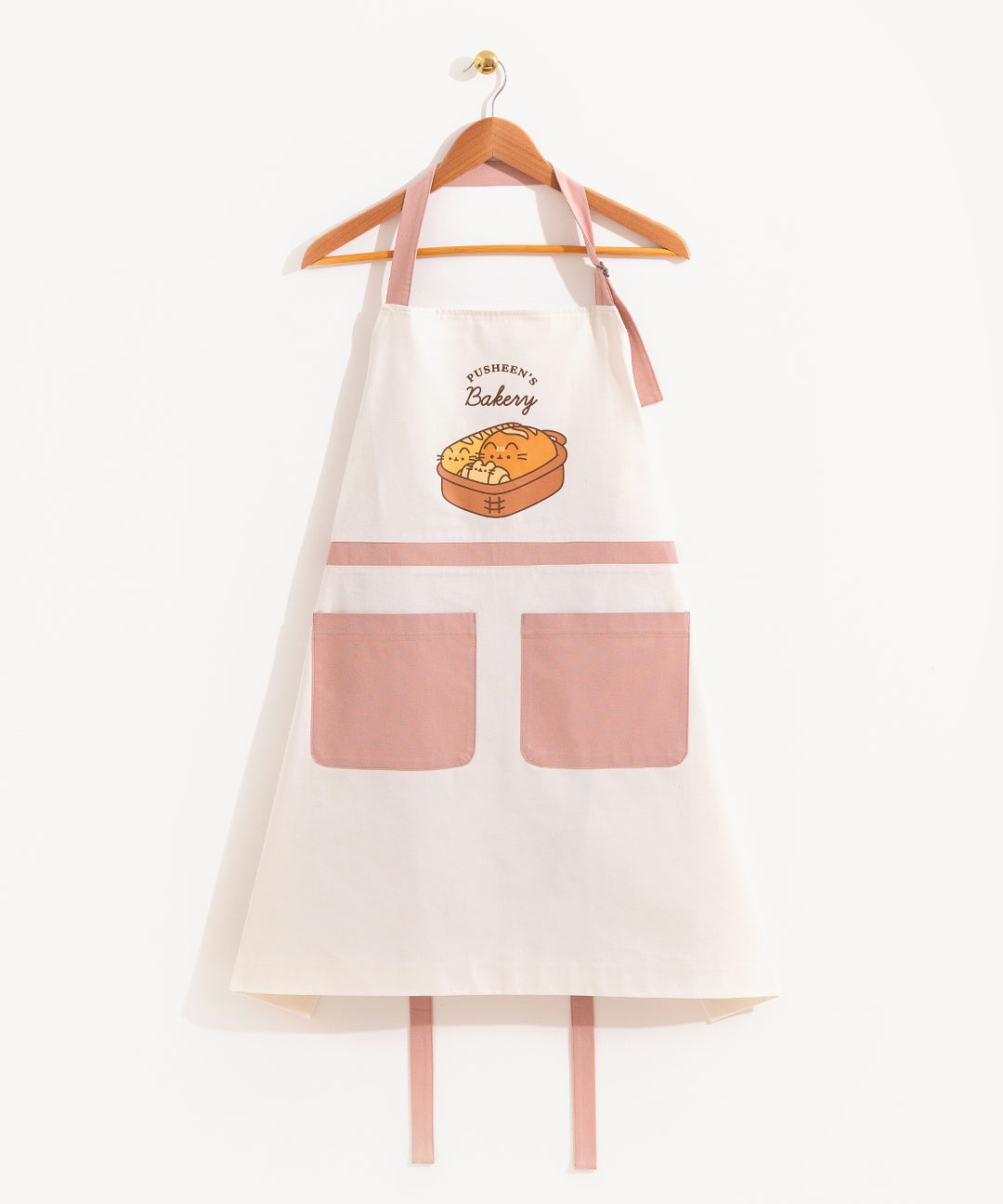 The Pusheen Breads Apron hanging from a clothes hanger in front of a light background. The neck strap has a g hook that allows it’s length to be adjusted. The apron draps off the hanger in the shape of a triangle, with the back straps dangling at the bottom behind the apron.