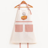 The Pusheen Breads Apron hanging from a clothes hanger in front of a light background. The neck strap has a g hook that allows it’s length to be adjusted. The apron draps off the hanger in the shape of a triangle, with the back straps dangling at the bottom behind the apron.