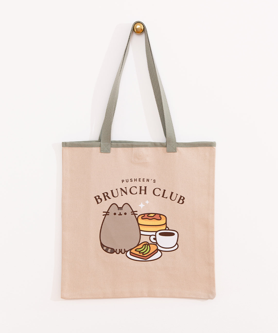 Pusheen Brunch Club Tote Bag hangs on a gold hook. The cream color cotton canvas bag has a sage green piping details and straps. There is a large Pusheen graphic printed on the front center of the tote.