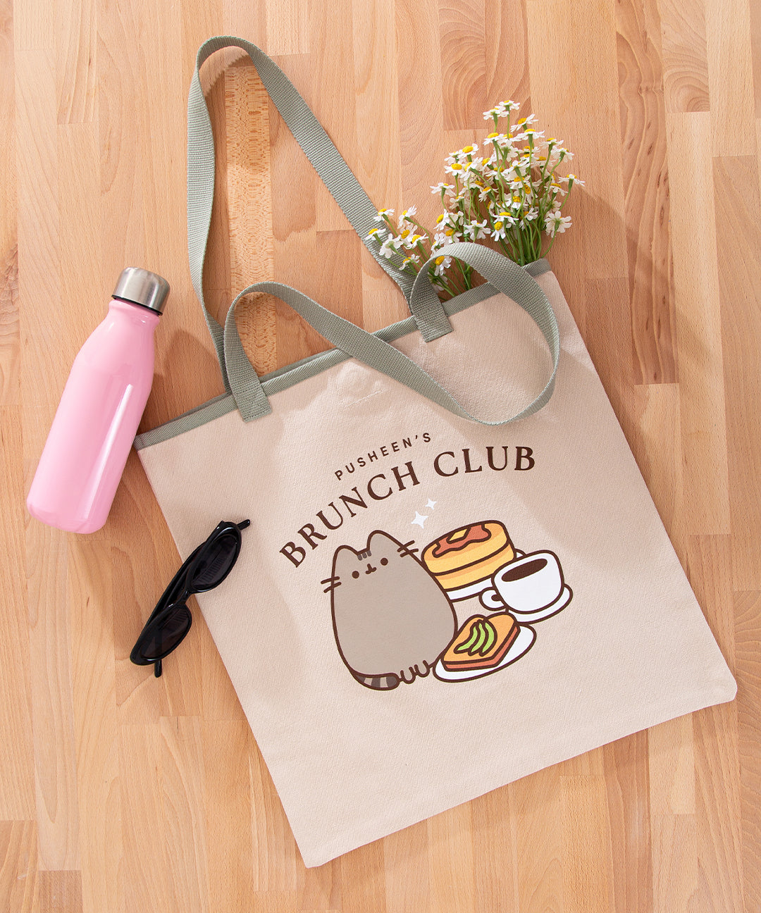 The cream and sage green Pusheen Brunch Club Tote Bag lies on a wooden surface surrounded by a pink water bottle, black sunglasses, and yellow and white daisies.
