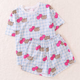 Cherry Pajama Set lies as a set on a white fluffy background. The oversized blue and white gingham print t-shirt has Pusheen the Cat and dark pink cherry graphics on the top. The stretchy blue and white shorts have the same pattern as the Pusheen pajama top. 