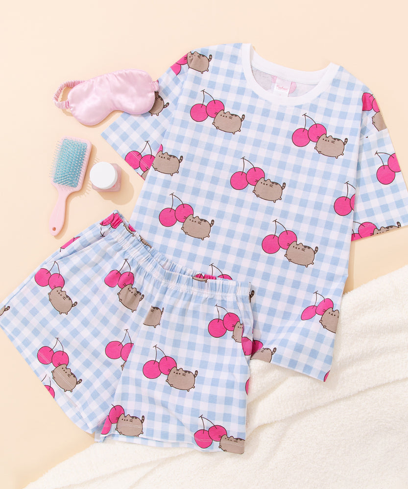 Pusheen Cherry Pajama Set includes a white and blue gingham oversized top and elastic waistband shorts. The set has an all over print of Pusheen the Cat leaping next to two dark pink cherries. 