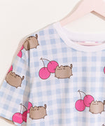 A close-up view of the white and blue pajama top. The top has an oversized fit and an all over print of Pusheen leaping next to two pink cherries. 