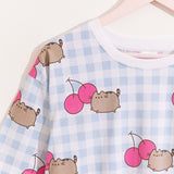 A close-up view of the white and blue pajama top. The top has an oversized fit and an all over print of Pusheen leaping next to two pink cherries. 