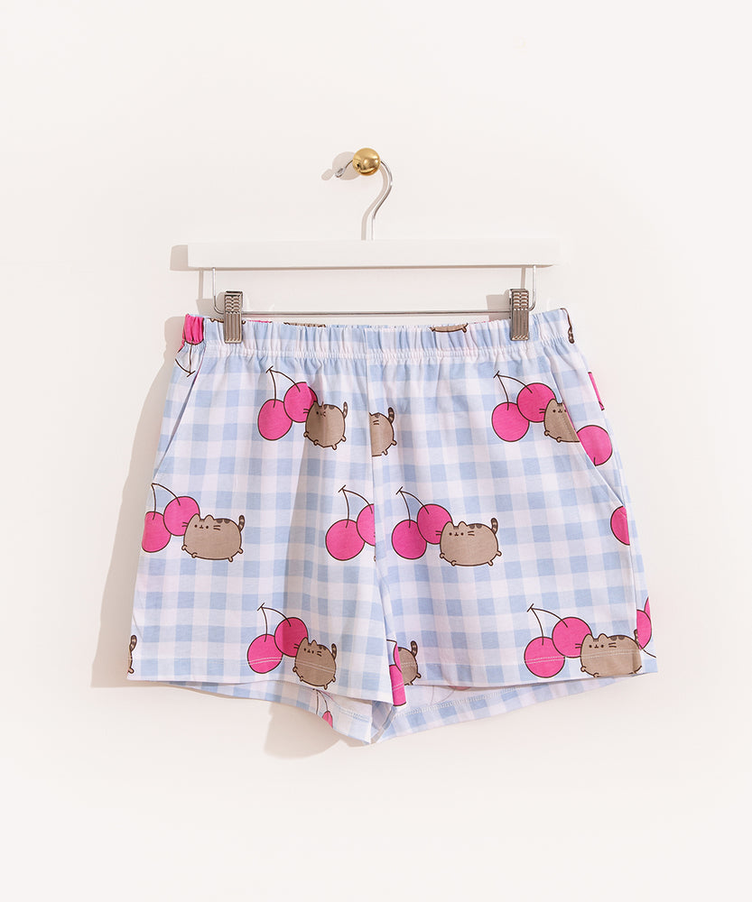 Hung on a white hanger, the Pusheen Pajama Shorts are hung to show the full length of the shorts. The blue and white shorts are covered in a pattern of gray Pusheens and dark pink cherries. 