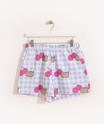 Hung on a white hanger, the Pusheen Pajama Shorts are hung to show the full length of the shorts. The blue and white shorts are covered in a pattern of gray Pusheens and dark pink cherries. 