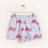 Hung on a white hanger, the Pusheen Pajama Shorts are hung to show the full length of the shorts. The blue and white shorts are covered in a pattern of gray Pusheens and dark pink cherries. 