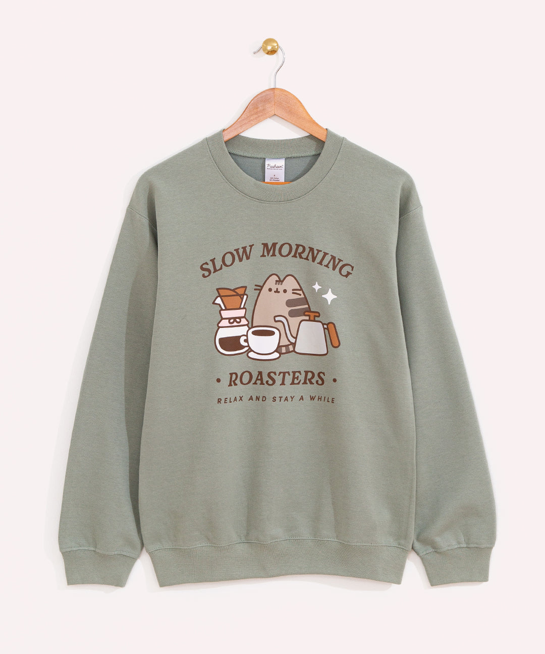Pusheen Coffee Sweatshirt hangs on a wooden hanger. The sage green sweatshirt shows Pusheen the cat surrounded by coffee and accessories.