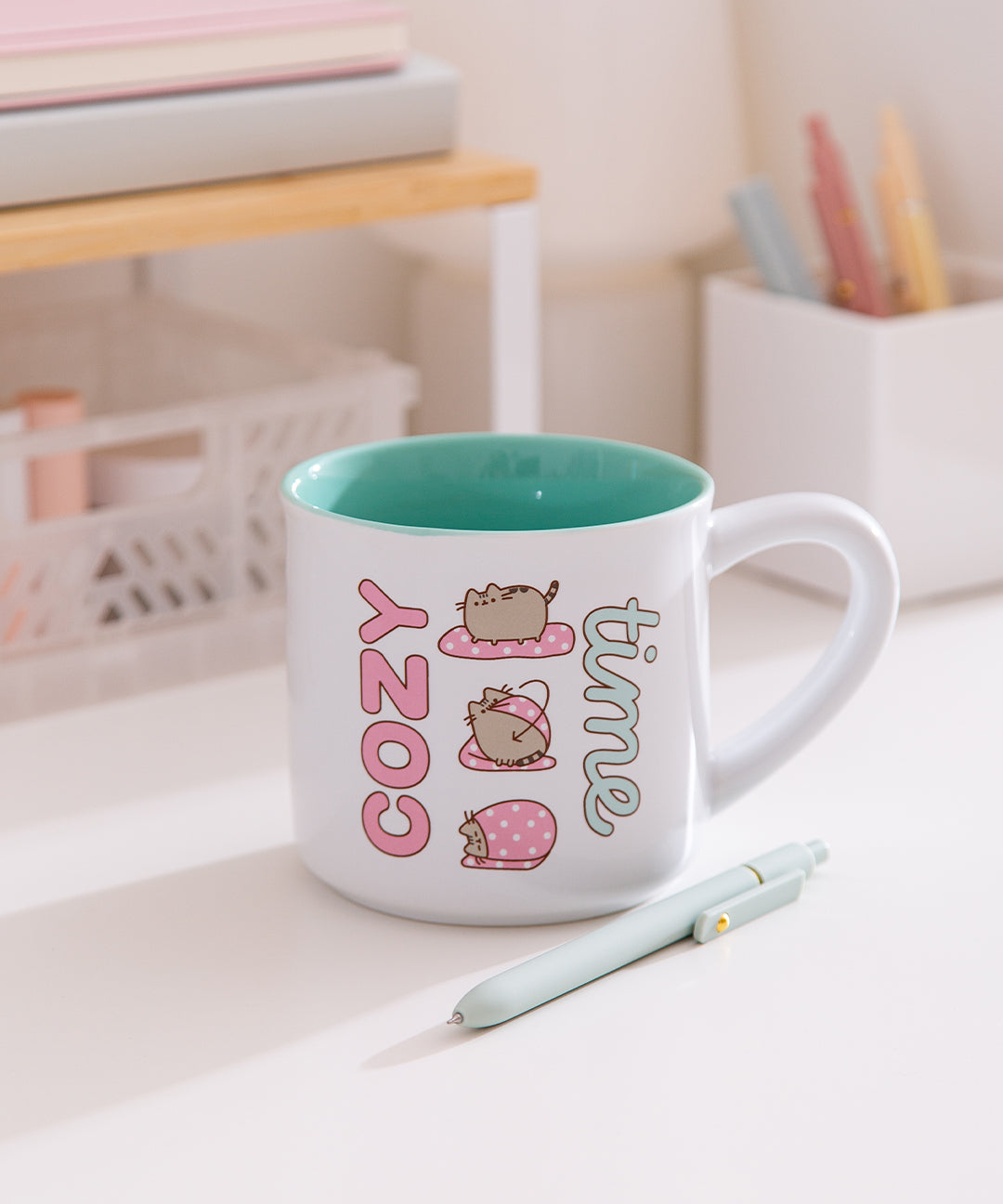 Pusheen Cozy Time Mug sits on a white desktop. The white mug has a mint green interior. The outside has a printed graphic of Pusheen the Cat wrapping herself in a blanket.