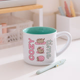 Pusheen Cozy Time Mug sits on a white desktop. The white mug has a mint green interior. The outside has a printed graphic of Pusheen the Cat wrapping herself in a blanket.