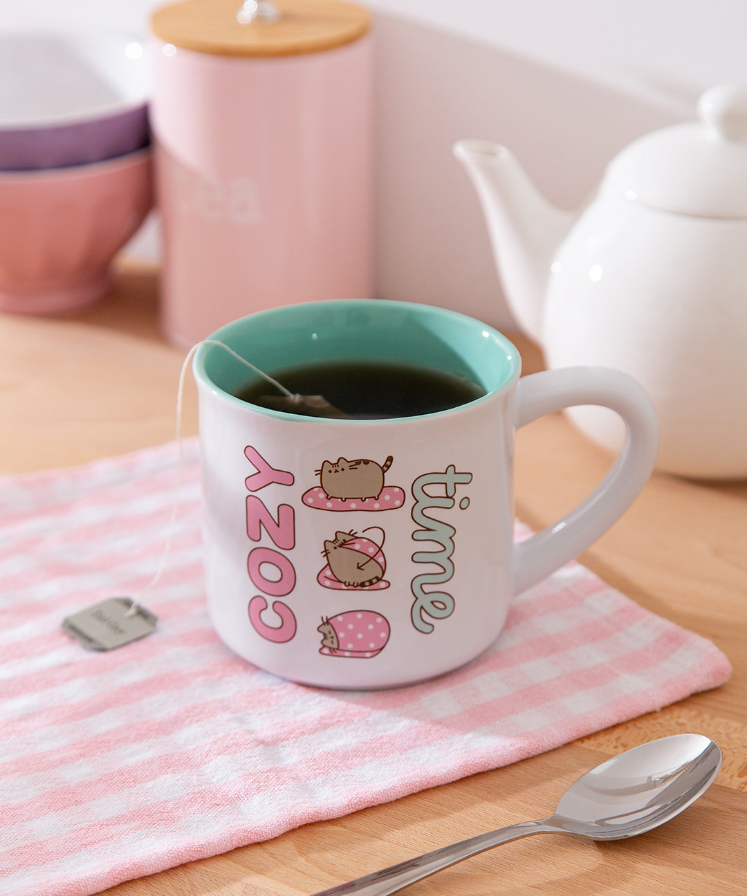 White mug with 'cozy time' text and Pusheen purrito graphic sits on a wooden table with tea and teapot.