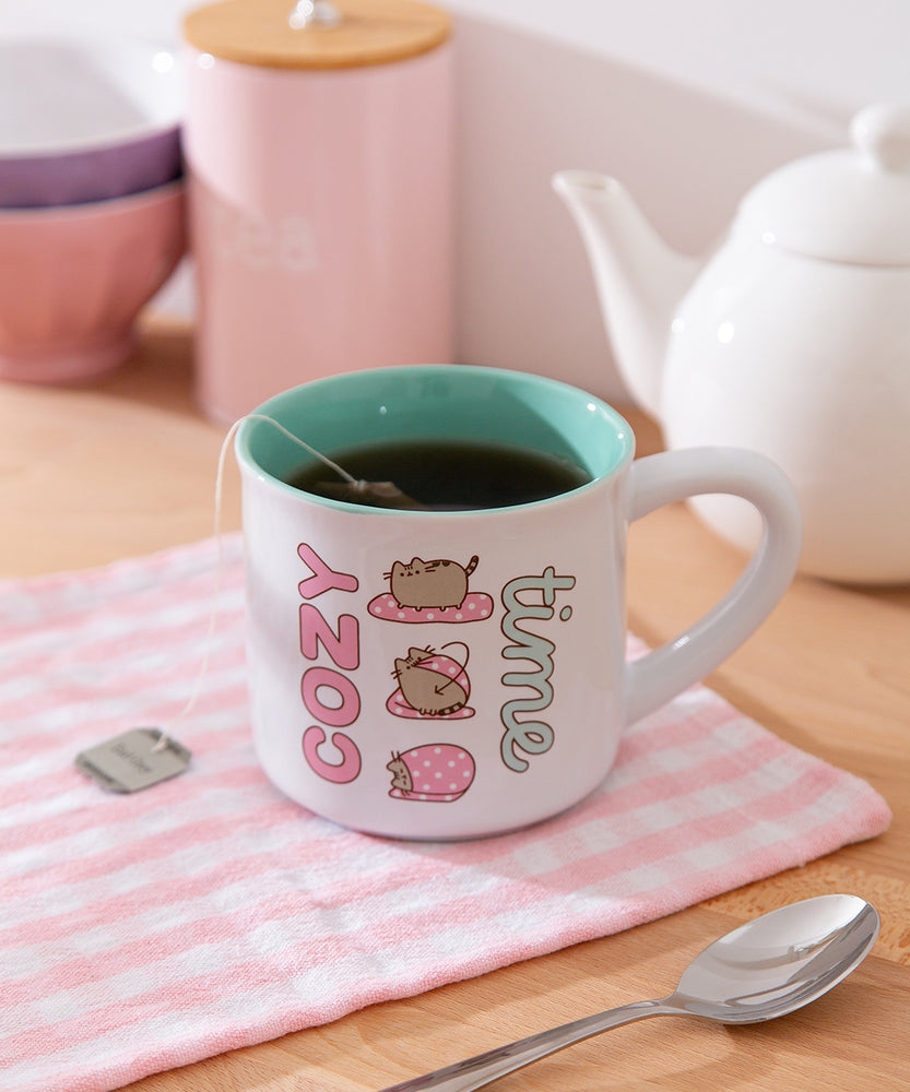 Ceramic Pusheen Mug sits on a brown desktop. The mug features Pusheen designs on each side of the mug.