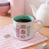 White mug with 'cozy time' text and Pusheen purrito graphic sits on a wooden table with tea and teapot.