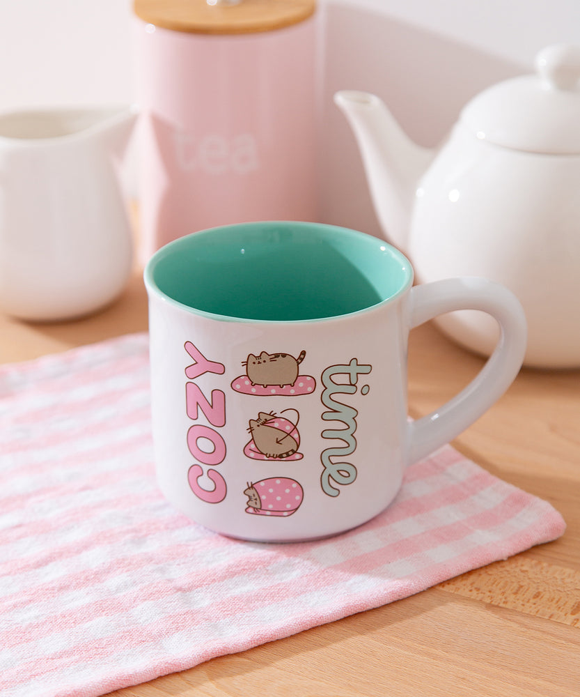 White mug with mint green interior has graphics printed on the exterior of Pusheen the Cat wrapping herself in a pink polka dot blanket. 