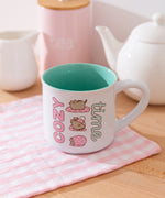 White mug with mint green interior has graphics printed on the exterior of Pusheen the Cat wrapping herself in a pink polka dot blanket. 