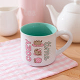 White mug with mint green interior has graphics printed on the exterior of Pusheen the Cat wrapping herself in a pink polka dot blanket. 