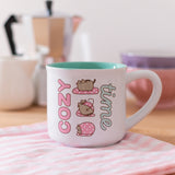 Close-up of the printed pink, white, grey, brown, and mint green graphic on the Pusheen Cozy Time mug that has a white exterior and green interior.