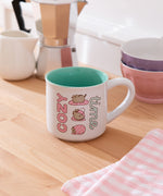 Ceramic Pusheen Mug sits on a brown desktop. The mug features Pusheen designs on each side of the mug.