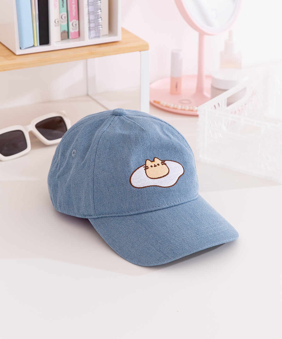 Top-down view of the Pusheen Denim Cap. The top button and seams can be seen from this view as well as the thread detailing on the bill of the hat. 
