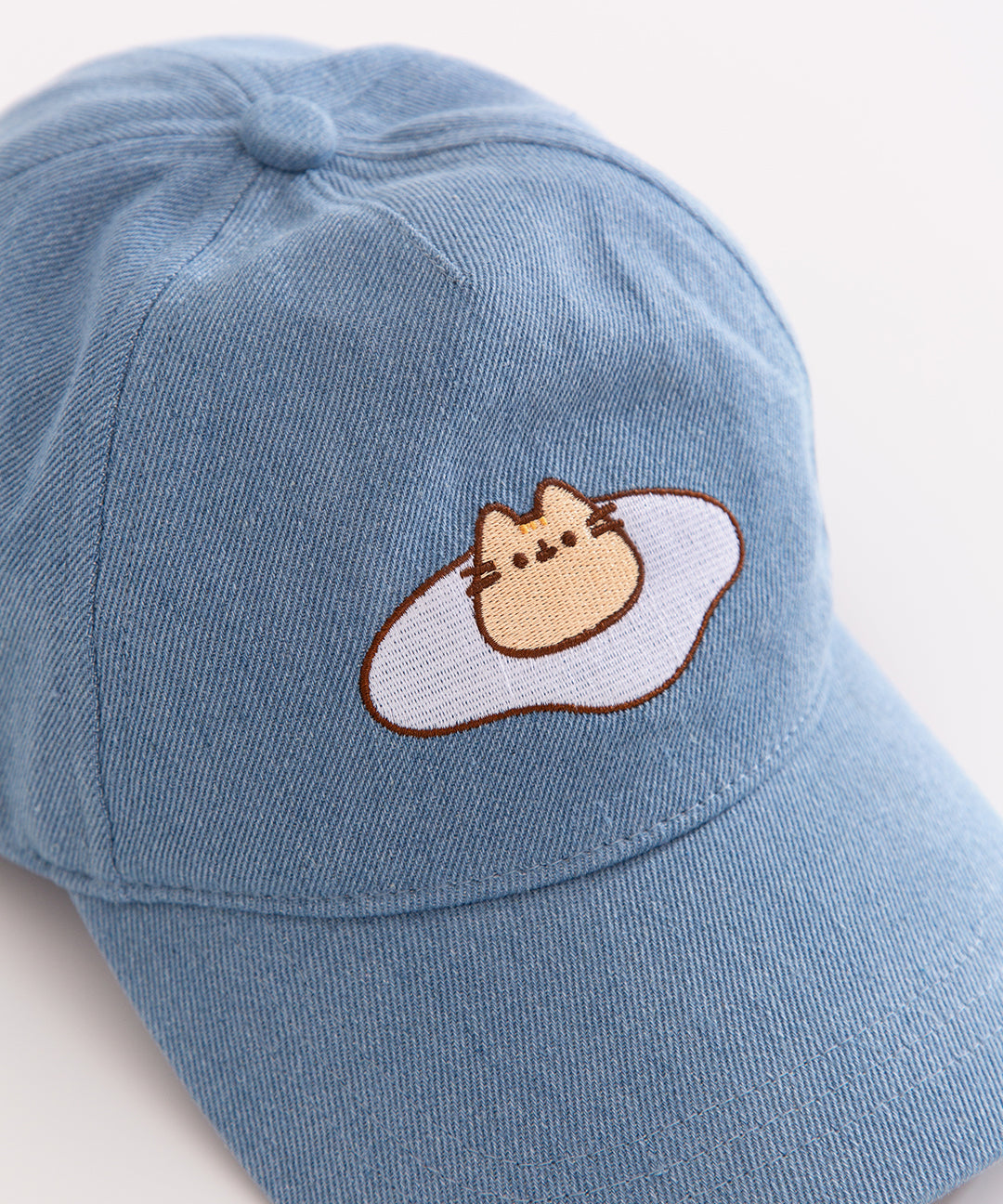 Close up view of embroidered graphic on the front center of the baseball style cap. Pusheen the Cat’s is outlined in dark brown and her ears, eyes, and mouth are shown in same dark brown thread. Pusheen's head stripes are dark yellow.
