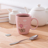 Pink mug holds tea and a tea bag while sitting on a desktop. The pink mug has a graphic of a gray tabby cat.
