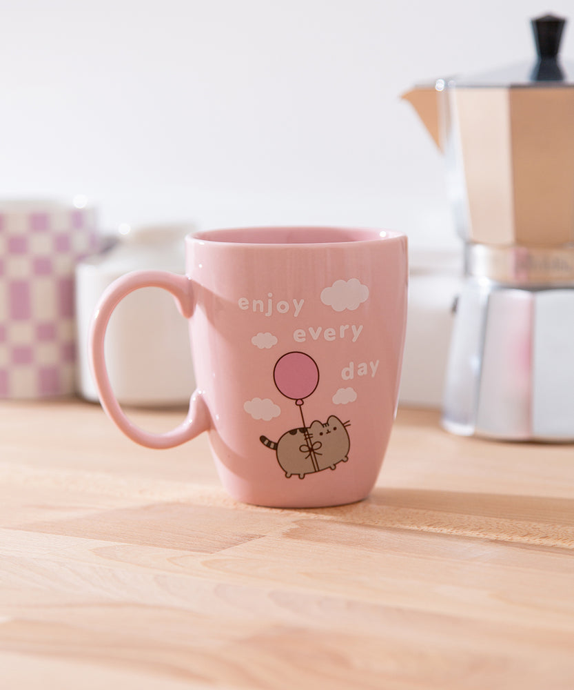 Pink Pusheen Enjoy Every Day Mug shows Pusheen the Cat floating in the white fluffy clouds with the phrase 'enjoy every day'.