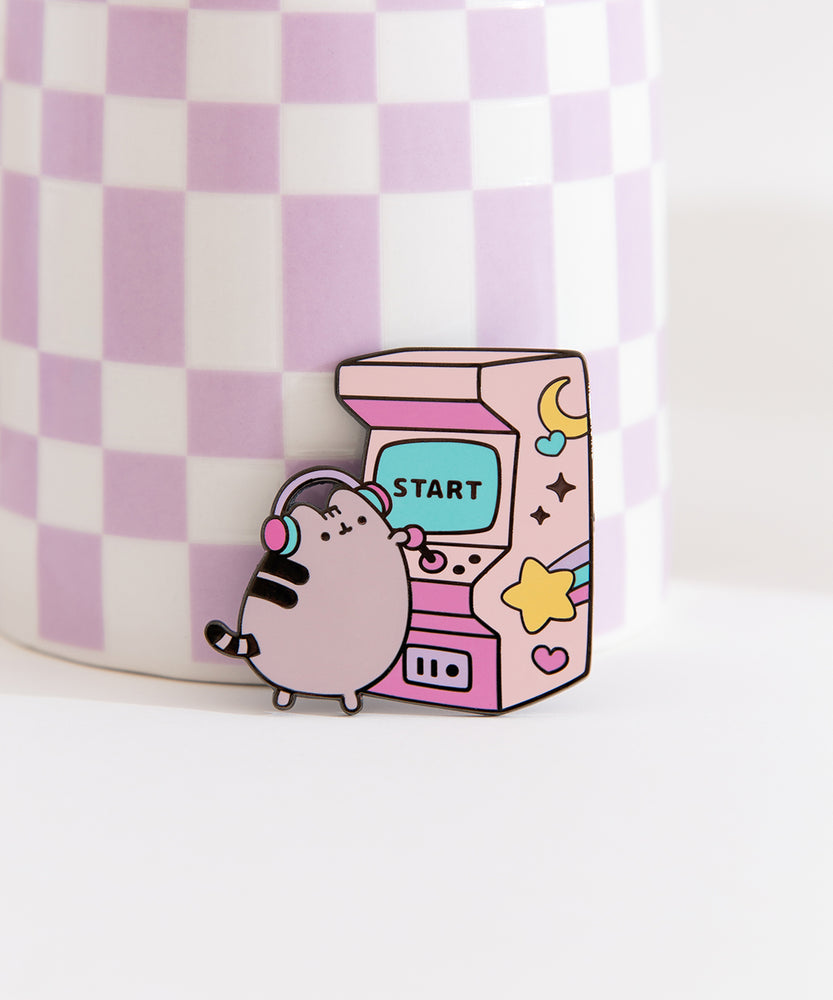 Close-up of Pusheen gamer pin. A gray cat wearing purple, pink, and blue headphones plays a classic arcade game. The large gaming console has a blue screen that says "start."
