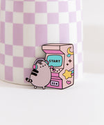 Close-up of Pusheen gamer pin. A gray cat wearing purple, pink, and blue headphones plays a classic arcade game. The large gaming console has a blue screen that says "start."