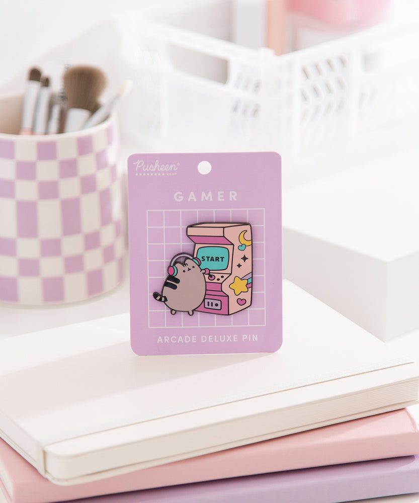 Front view of the Pusheen Gamer Arcade Deluxe Pin on its purple backer card. A grey cat stands to play a pink retro arcade game. 