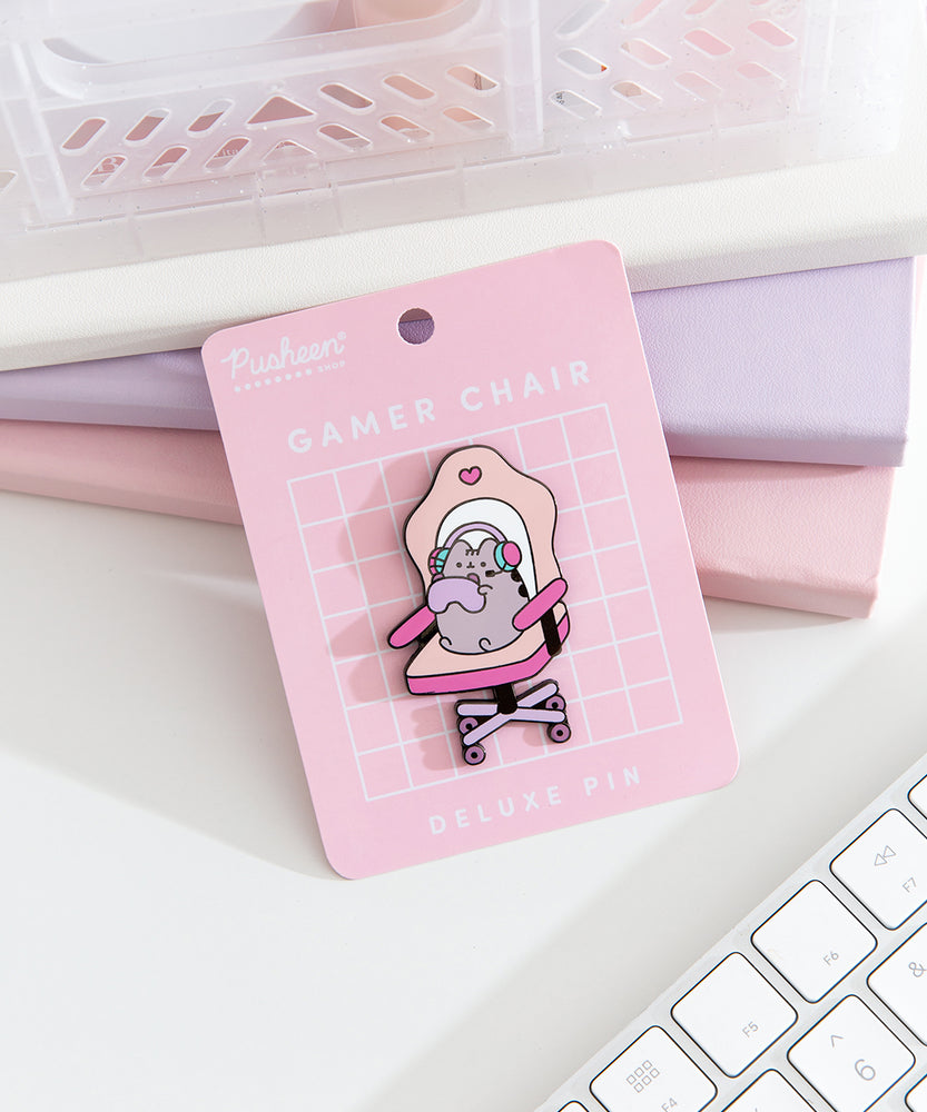 Front view of the Pusheen Gamer Chair Deluxe Pin on its pink backer card. A grey cat sits in an oversized chair to play online games.