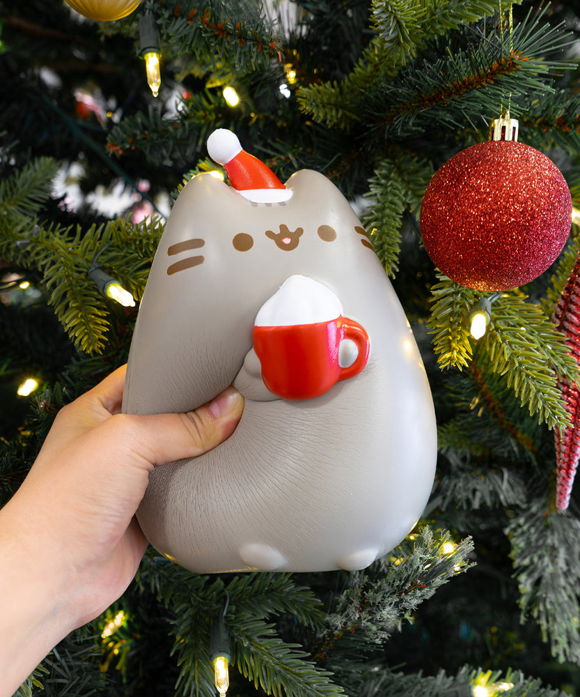 Front view of the Pusheen Holiday Jumbo Squishy. The gray cat is wearing a white and red santa hat while holding a red mug with white whipped cream.