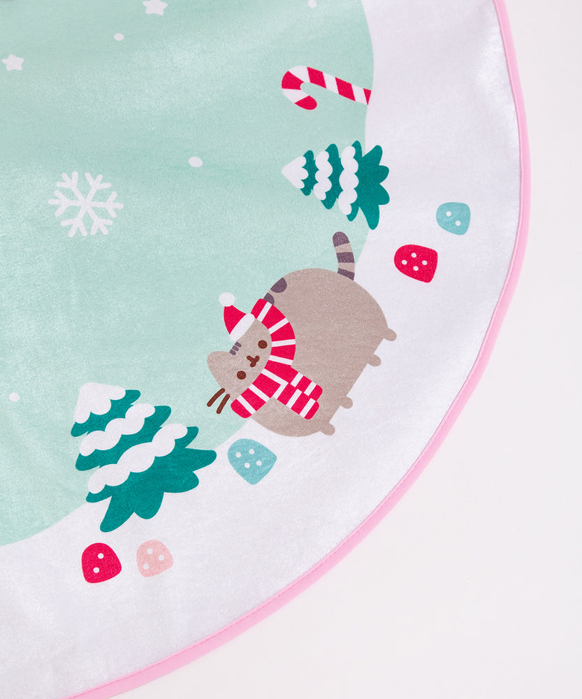 Close-up of the snowy landscape print on the tree skirt. Pusheen is shown wearing a Santa Hat next to two snowcapped pine trees.