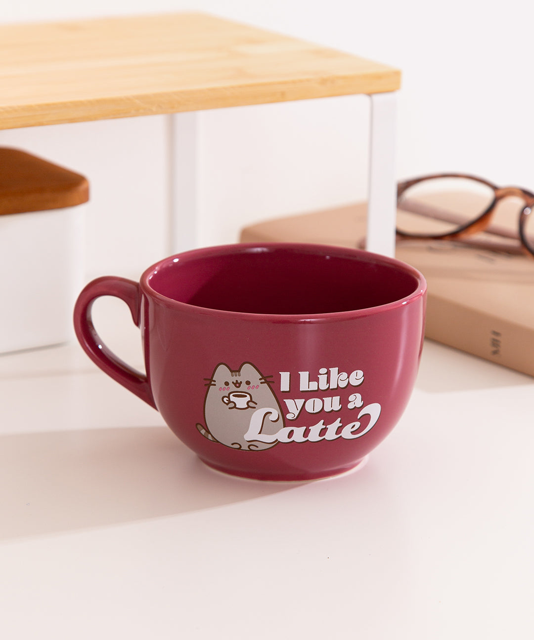 Pusheen I Like You a Latte ceramic mug sits on a tabletop. The red mug has a graphic of Pusheen the Cat blushing and holding a hot beverage in her paws.