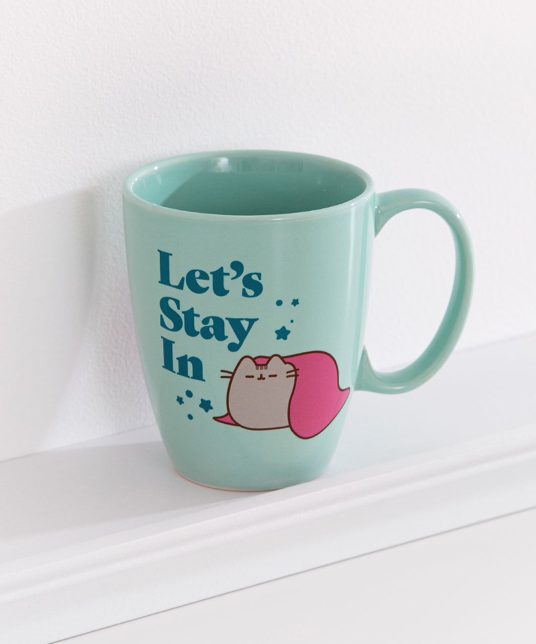 Pusheen Let's Stay In ceramic mug is light green and features a graphic of a gray tabby cat resting under a blanket surrounded by stars and the phrase "Let's Stay In."