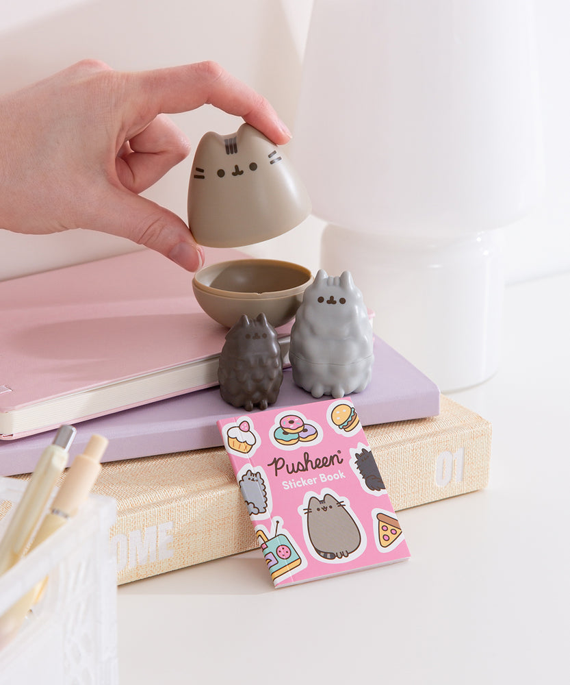 Pusheen Mini Nesting Dolls set featuring Pusheen, Stormy, and Pip sit in front of the packaging box that the dolls come in and the included sticker book.
