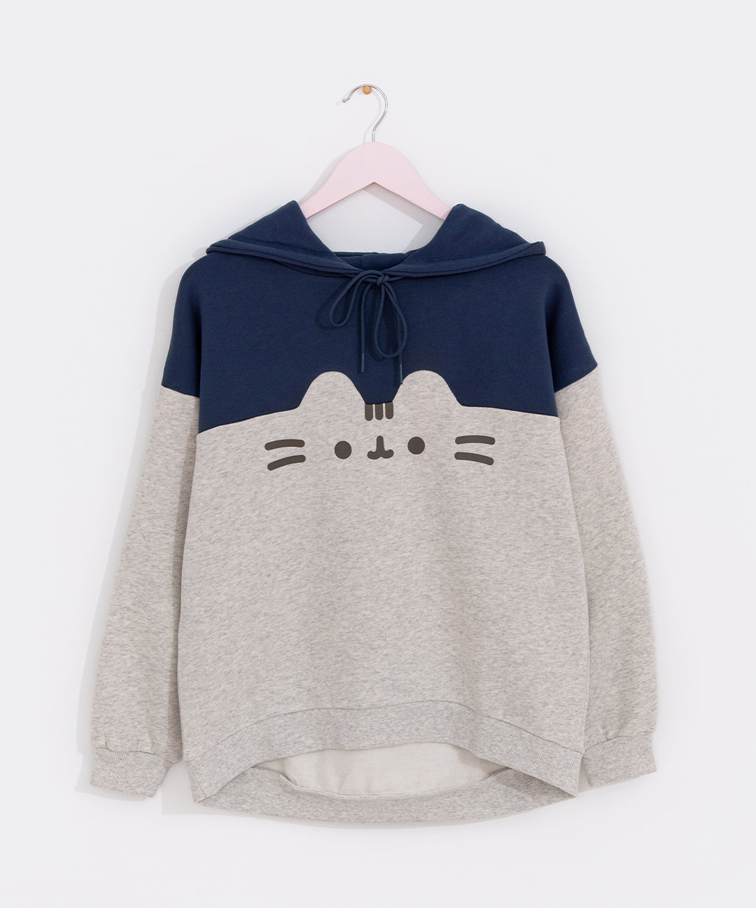Pusheen Navy Relaxed-Fit Character Hoodie hangs on a light pink hanger in front of a white wall. The two-toned sweatshirt features Pusheen’s face in the center of the hoodie. The top half of the hoodie is navy blue while the bottom portion is light gray and outlines Pusheen’s rounded triangle ears to separate the two colors. 
