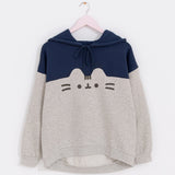 Pusheen Navy Relaxed-Fit Character Hoodie hangs on a light pink hanger in front of a white wall. The two-toned sweatshirt features Pusheen’s face in the center of the hoodie. The top half of the hoodie is navy blue while the bottom portion is light gray and outlines Pusheen’s rounded triangle ears to separate the two colors. 