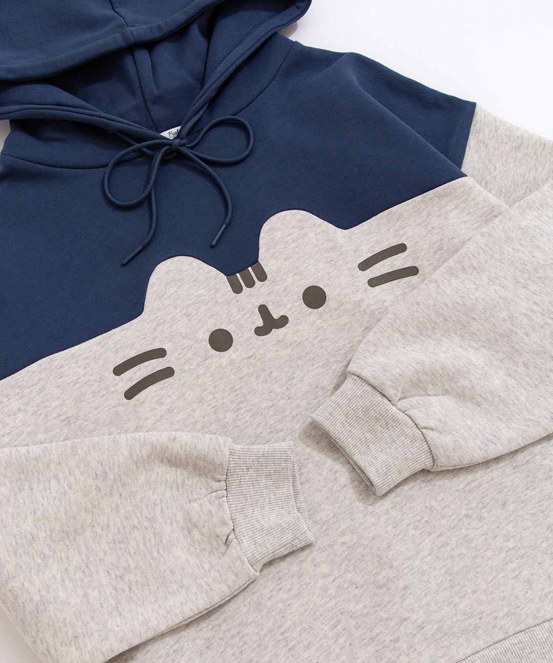 A close-up view of the printed graphic on the front center chest of the sweatshirt. The classic Pusheen pose shows her whiskers, mouth, eyes,head stripes in a dark gray print. The navy blue hoodie drawstring is tied in a bow.  The sleeves and cuffs are folded and shown.