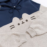A close-up view of the printed graphic on the front center chest of the sweatshirt. The classic Pusheen pose shows her whiskers, mouth, eyes,head stripes in a dark gray print. The navy blue hoodie drawstring is tied in a bow.  The sleeves and cuffs are folded and shown.
