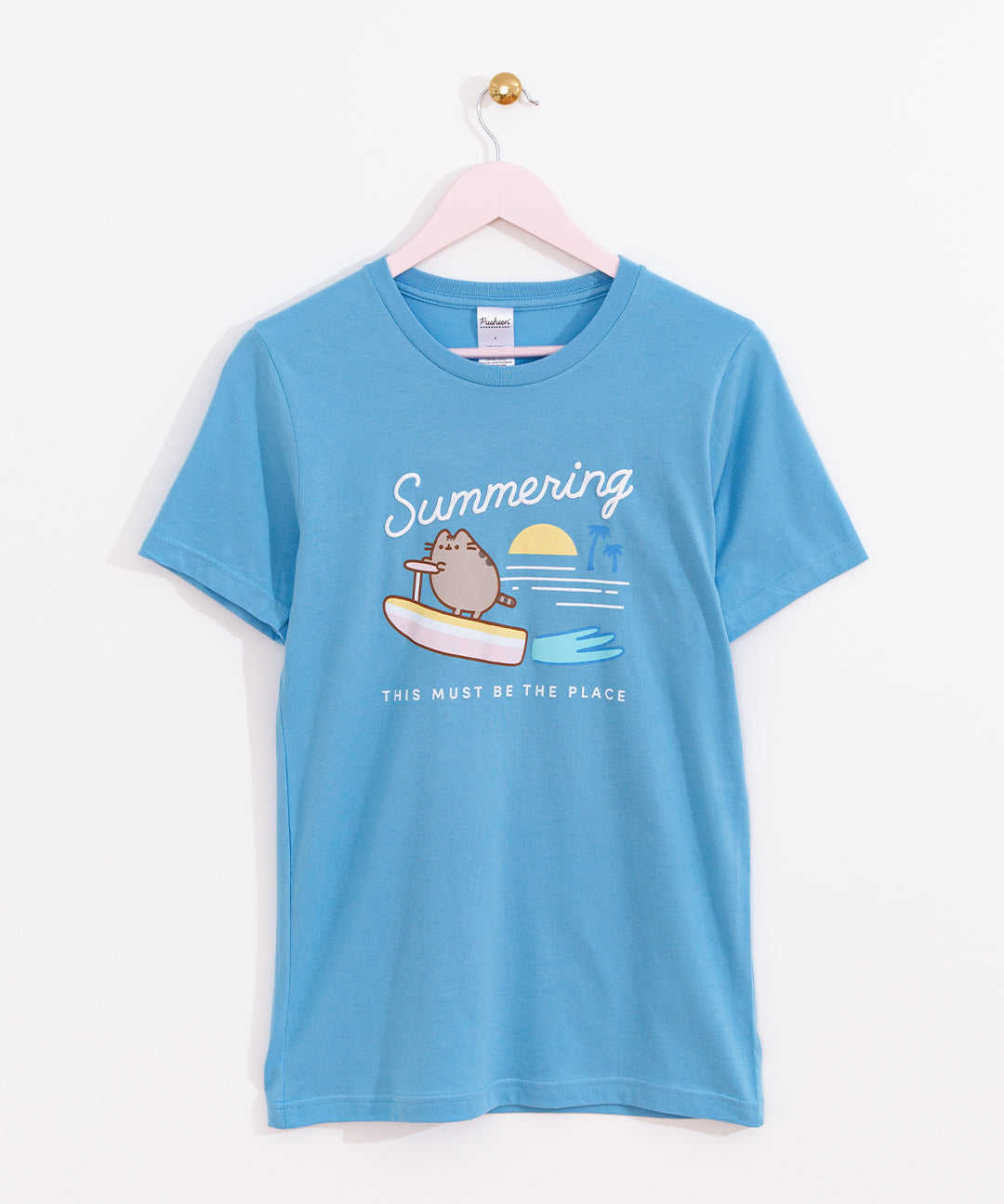 Blue Pusheen Tee-Shirt shows Pusheen the Cat "summering" while riding on a surfboard in the ocean.