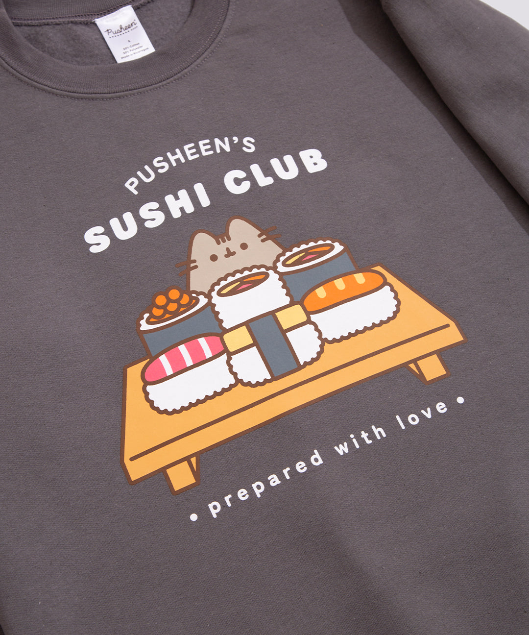 Close-up of front graphic on the charcoal-colored Pusheen sweatshirt. Above the graphic of Pusheen with a selection of sushi is the phrase "Pusheen's Sushi Club" in a white print. Beneath the graphic of different fish sushi rolls is the phrase "prepared with love"