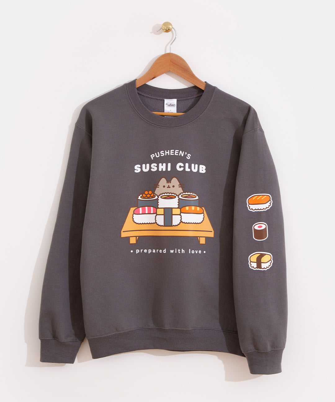 Pusheen Sushi Club Sweatshirt hangs on a brown hanger in front of a white wall. The charcoal grey sweatshirt features a large graphic of Pusheen with sushi pieces and rolls on the front center of the sweatshirt. There is a print down the wearer's left sleeve of sushi icons.