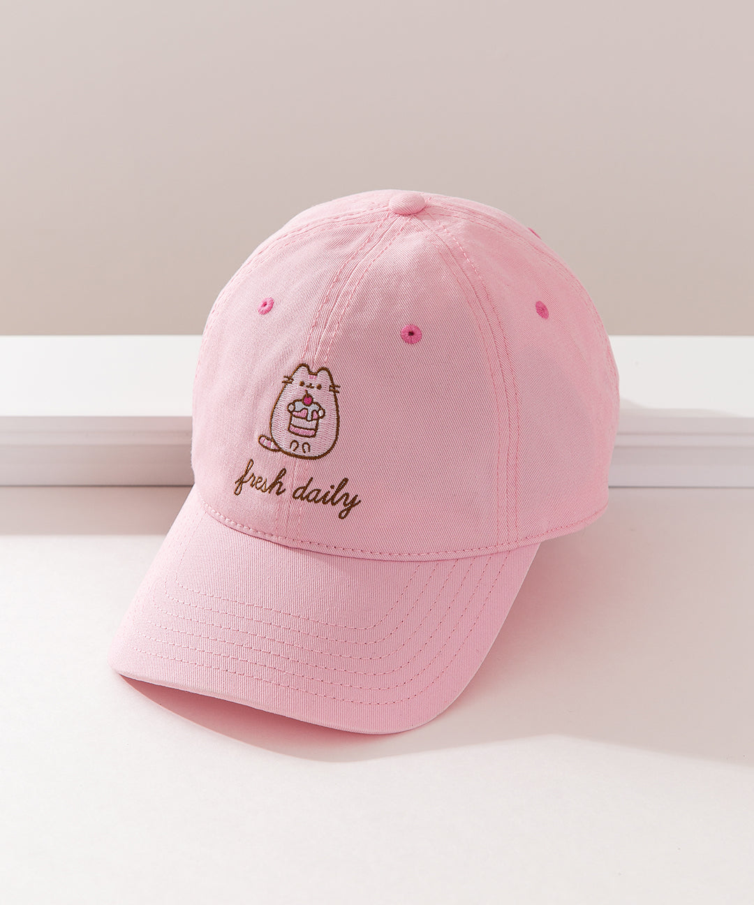 Top-down view of the Pusheen Pink Cap. The light pink dad hat has an embroidery of Pusheen holding a cherry dessert that will show on the wearer's forehead area. 
