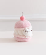 Side quarter view of the cherry macaron Pusheen plush. The macaron plush has a light pink shells with a white whipped filling in the middle. 