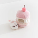 Pink plush toy resembling a macaron with a small white Pusheen the cat sitting next to the macaron plush on a light background.