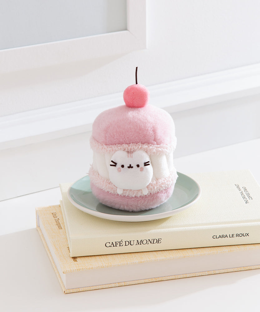 Pink plush toy resembling a macaron with a cherry on top, sitting on a stack of books.