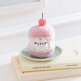 Pink plush toy resembling a macaron with a cherry on top, sitting on a stack of books.