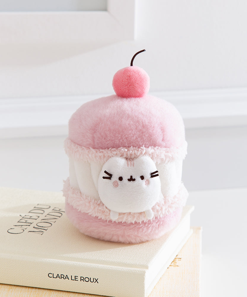 Side quarter view of the cherry macaron Pusheen plush. The macaron plush has a light pink shells with a white whipped filling in the middle. 