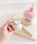 Model holds the removable Pusheen the Cat white plush that sits inside the cherry macaron plush environment. The light plush rests between the model’s fingers.  